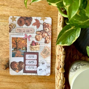 Puede incluir: Una tarjeta decorativa con un collage de pegatinas con temática otoñal. La tarjeta incluye un oso de peluche, manzanas, una escena de árbol y el texto "Today I'd like to sit and read". También se presentan entradas para el "Late Night Book Club" y pegatinas de fantasmas.