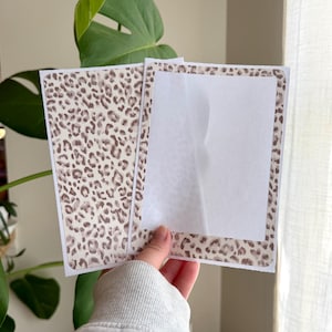 May include: Two rectangular paper cards featuring a brown and beige leopard print pattern. One card displays the full animal print design, while the other card features a leopard print border surrounding a large, blank, white rectangular center area.