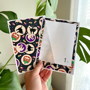 May include: Two rectangular sticker sheets featuring a black background with a repeating pattern of Halloween-themed cookies, including black cats, ghosts, and pumpkins, interspersed with candy corn. One sheet has a white backing labeled 1.