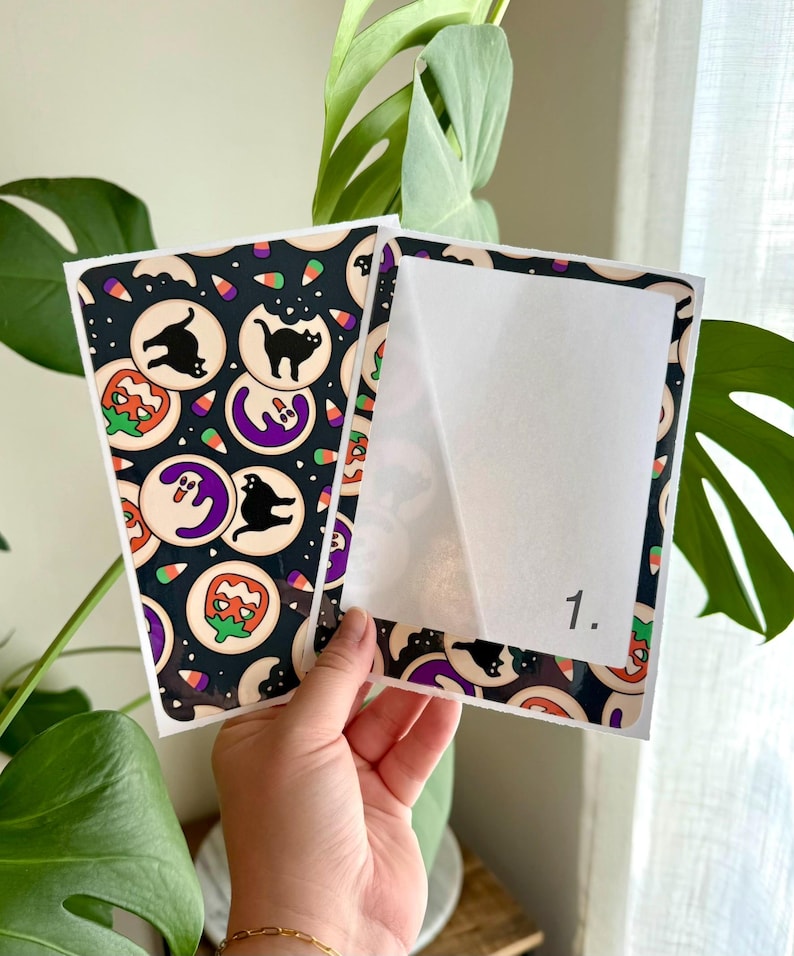May include: Two rectangular sticker sheets featuring a black background with a repeating pattern of Halloween-themed cookies, including black cats, ghosts, and pumpkins, interspersed with candy corn. One sheet has a white backing labeled 1.