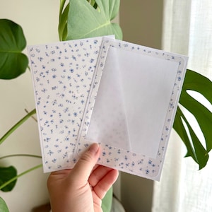 Puede incluir: Una tarjeta rectangular blanca con un delicado patrón floral azul en su superficie. La tarjeta incluye un marco rectangular central con una capa blanca translúcida, bordeado por el mismo diseño floral azul sobre un fondo blanco.