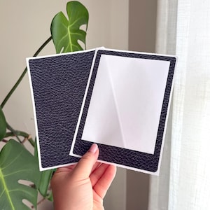 May include: Two rectangular cards featuring a dark navy blue textured faux leather finish. One card is solid, while the other has a white rectangular center cutout framed by the same dark navy blue textured material, resembling a classic photo frame.