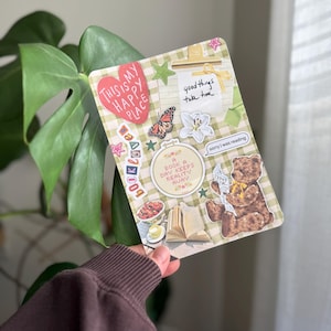 Puede incluir: Una tarjeta decorativa con un patrón de cuadros verdes y blancos, adornada con pegatinas. Las pegatinas incluyen un corazón que dice "This is my happy place", una mariposa, un oso de peluche leyendo un libro y el texto "good things take time".