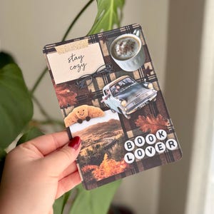 Puede incluir: Una tarjeta rectangular con un fondo de cuadros marrón oscuro y elementos de collage que incluyen una taza de café, gafas negras, un coche antiguo, un cruasán, un paisaje y el texto "stay cozy" y "BOOK LOVER" en letras blancas.