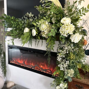 May include: A large floral arrangement featuring white hydrangea and peony blossoms mixed with various green foliage, ferns, and wispy grasses. The arrangement is draped across a horizontal surface, with one side cascading downward in a lush display.