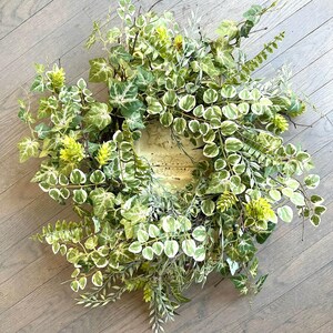 May include: A green and white leafy wreath with a natural woven base. The wreath is made of artificial greenery and has a rustic look.