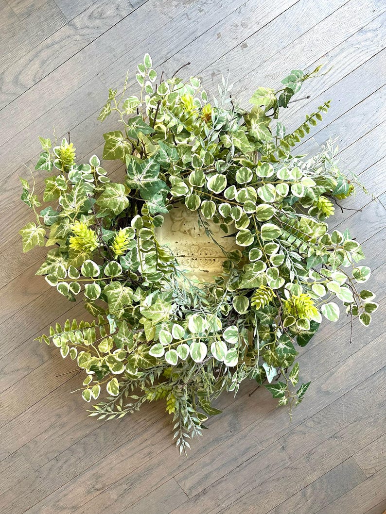 May include: A green and white leafy wreath with a natural woven base. The wreath is made of artificial greenery and has a rustic look.