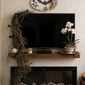May include: An oval decorative plate featuring a central bat illustration, mounted above a television. A wooden mantel holds a trailing vine with small bat cutouts, a white orchid in a ceramic pot, a small candle, and a metallic tray.