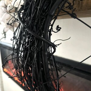 May include: A circular wreath constructed from thin, matte black intertwined twigs and vine tendrils. The structure features a dense, textured arrangement of dark, woody branches with small, curled offshoots extending outward from the main frame.