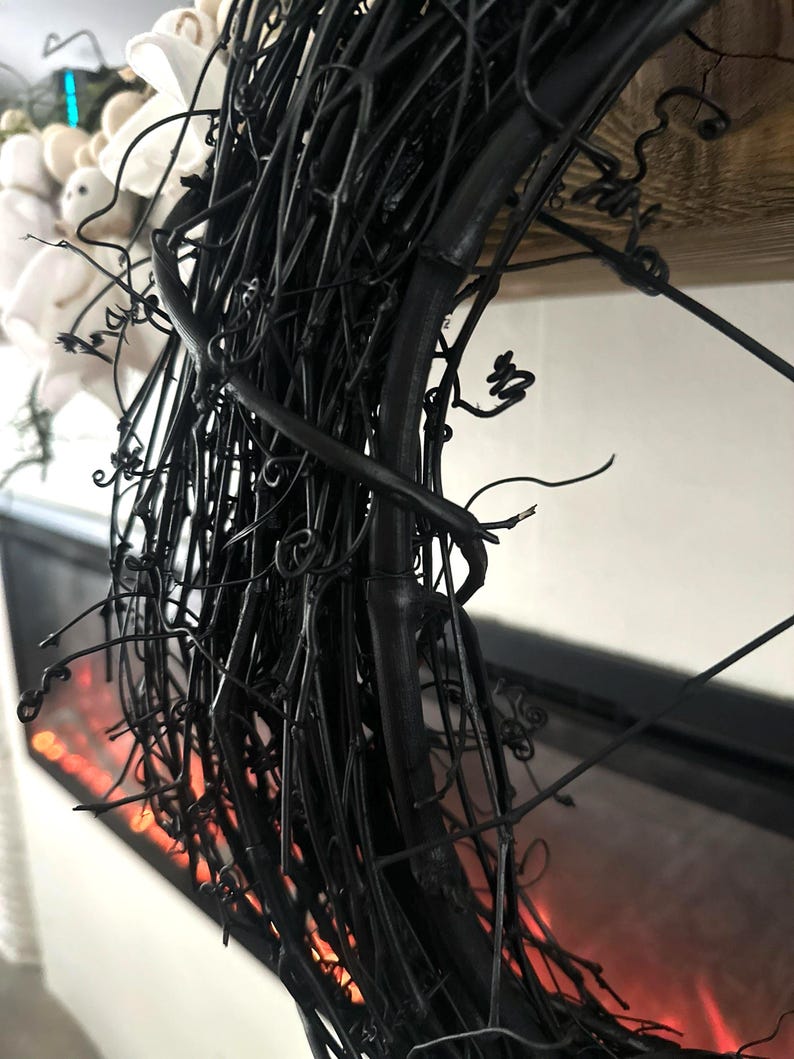 May include: A circular wreath constructed from thin, matte black intertwined twigs and vine tendrils. The structure features a dense, textured arrangement of dark, woody branches with small, curled offshoots extending outward from the main frame.