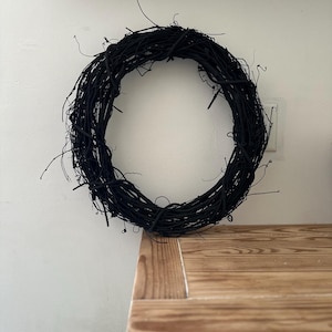 May include: A circular wreath constructed from thin, intertwined twigs and branches, all finished in a solid matte black color. The structure features a natural, organic texture with various small, protruding stems extending from the main ring.