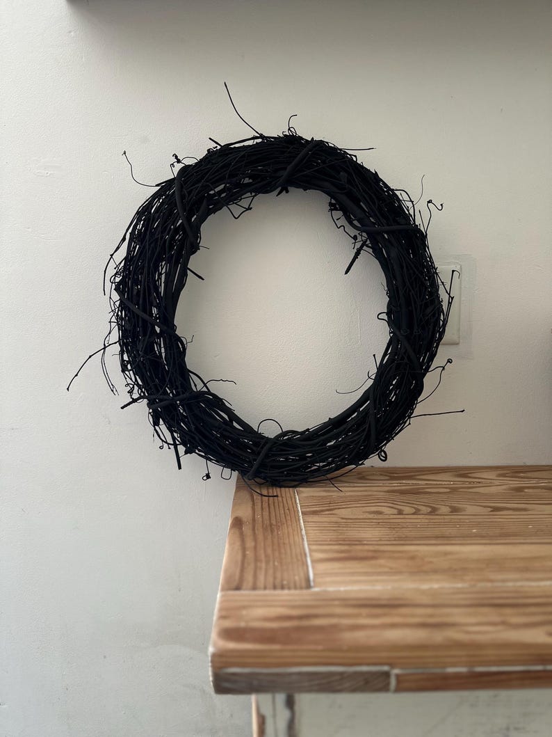May include: A circular wreath constructed from thin, intertwined twigs and branches, all finished in a solid matte black color. The structure features a natural, organic texture with various small, protruding stems extending from the main ring.