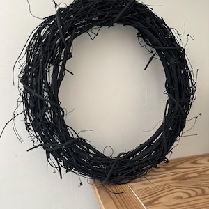 May include: A circular wreath constructed from intertwined, thin, matte black twigs. The structure features a dense, organic arrangement of branches with various protruding ends, creating a textured, uniform black appearance throughout the entire piece.
