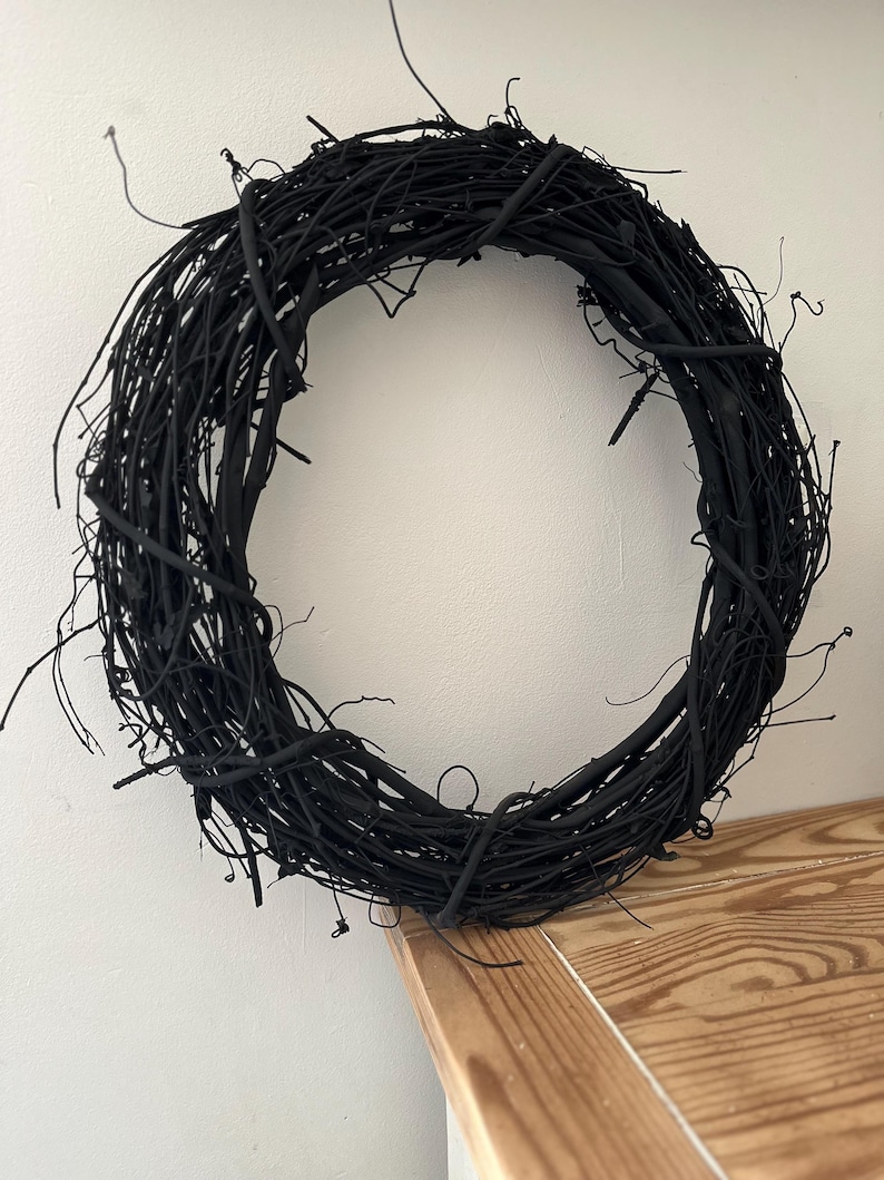 May include: A circular wreath constructed from intertwined, thin, matte black twigs. The structure features a dense, organic arrangement of branches with various protruding ends, creating a textured, uniform black appearance throughout the entire piece.