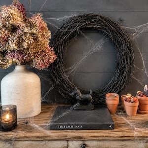 May include: A white ceramic vase filled with dried hydrangea flowers, a black twig wreath, a small black dog figurine, a book titled Tim Flach Dogs, three terracotta pots with dried flowers, and two dark glass candle holders with lit candles.
