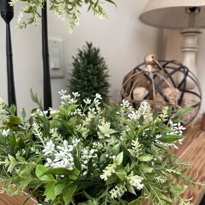 May include: A small green and white floral arrangement in a metal container. The arrangement is on a wooden surface with a lamp and other decor in the background.