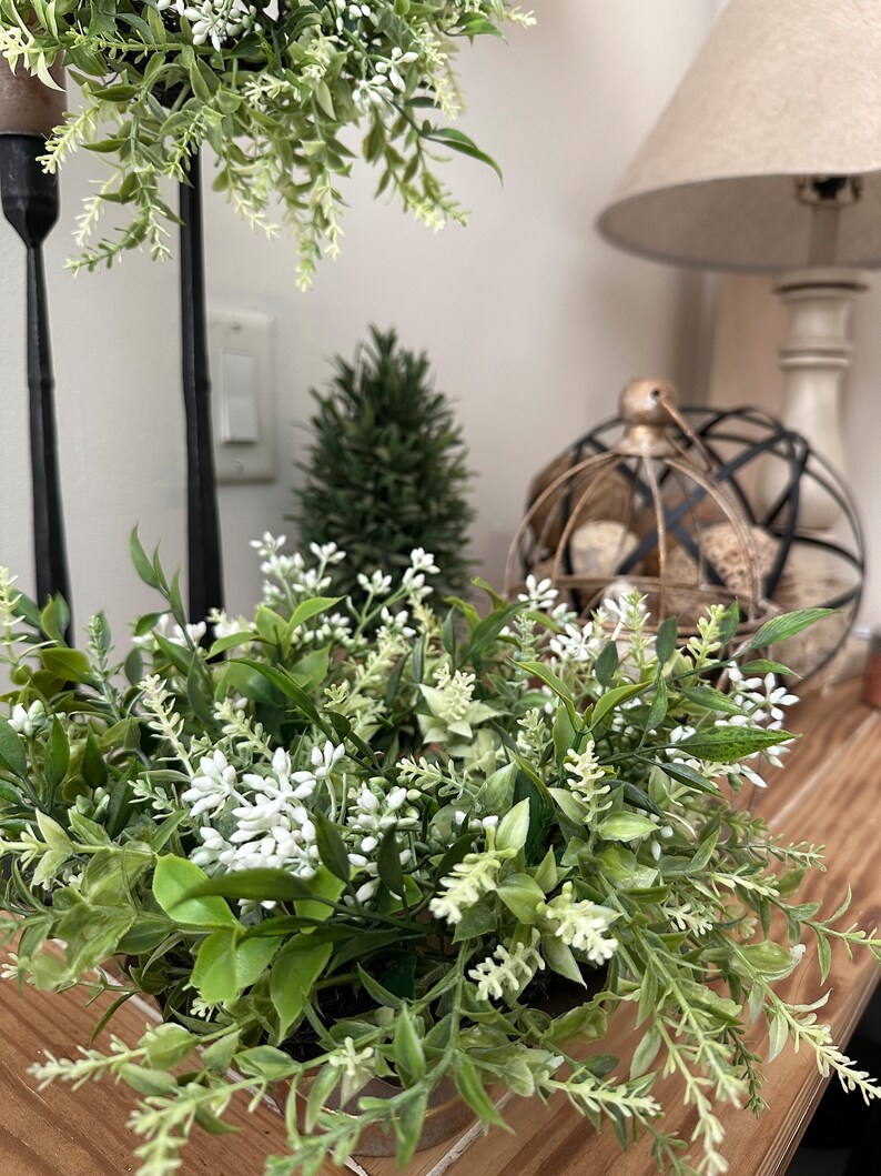 May include: A small green and white floral arrangement in a metal container. The arrangement is on a wooden surface with a lamp and other decor in the background.