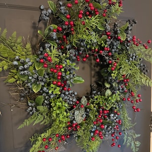 May include: A circular wreath constructed on a twig base, featuring a dense arrangement of green fern fronds, small green leaves, clusters of dark blue berries, and bright red berries distributed throughout the foliage in a balanced pattern.