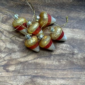 May include: A collection of six acorn-shaped ornaments featuring a gold-speckled top, a vibrant red band in the center, and a smooth white base. Each ornament is topped with a silver-colored cap and includes a thin gold hanging string.