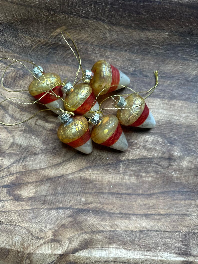 May include: A collection of six acorn-shaped ornaments featuring a gold-speckled top, a vibrant red band in the center, and a smooth white base. Each ornament is topped with a silver-colored cap and includes a thin gold hanging string.