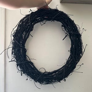 May include: A circular wreath constructed from intertwined, matte black twigs and branches. The structure features a dense, organic arrangement of thin, woody stems that form a continuous ring shape with various protruding, irregular twig ends.