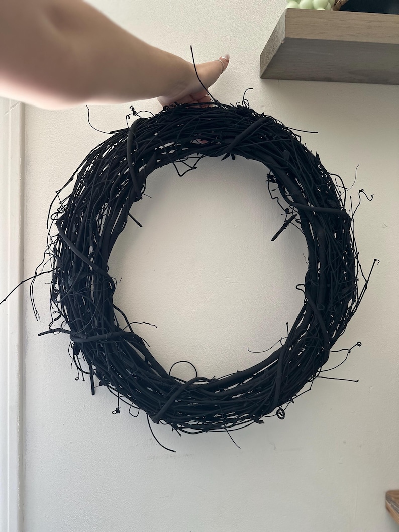 May include: A circular wreath constructed from intertwined, matte black twigs and branches. The structure features a dense, organic arrangement of thin, woody stems that form a continuous ring shape with various protruding, irregular twig ends.