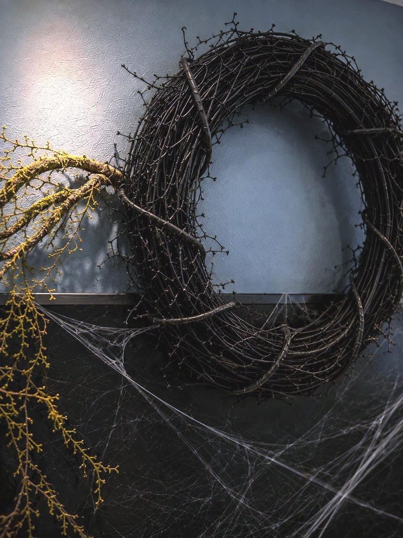 May include: A circular wreath constructed from dark brown, bare, thorny branches. A single, moss-covered branch extends outward from the left side of the wreath. The lower portion of the wreath is draped with thin, white, translucent spiderwebs.