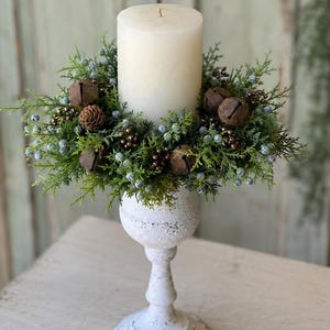 May include: A white pillar candle sits in a decorative wreath of green cedar foliage, small blue berries, brown pinecones, and rustic metal jingle bells, all supported by a white pedestal candle holder with a distressed, textured finish.