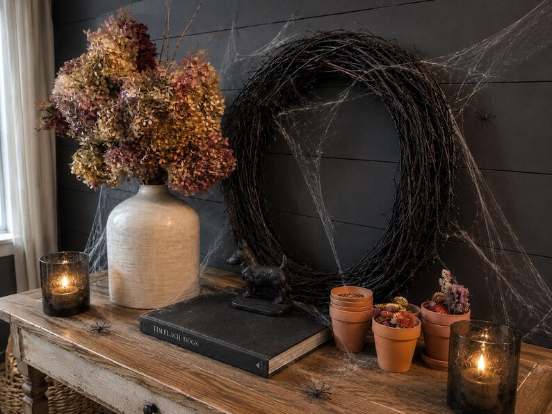 May include: A white ceramic vase with dried hydrangea flowers, a black twig wreath covered in artificial spiderwebs, a black dog figurine, a book titled Tim Flach Dogs, three terracotta pots with dried flowers, and two dark glass candle holders.