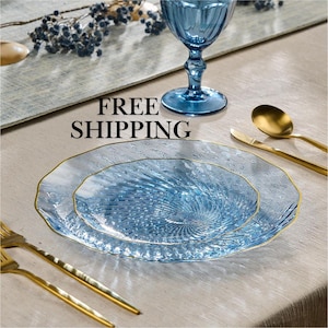 May include: A light blue glass dinner plate and a smaller salad plate, both featuring a textured spiral pattern and a thin gold rim. The set is accompanied by a blue glass goblet, a gold-toned spoon, and a gold-toned fork on a neutral fabric.