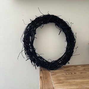 May include: A circular wreath constructed from thin, intertwined twigs and branches, entirely painted in a matte black finish. The structure features a dense, organic arrangement of woody stems with numerous small, protruding ends.