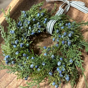 May include: A green wreath with blue berries and a white and black striped ribbon. The wreath is sitting in a wooden bowl.