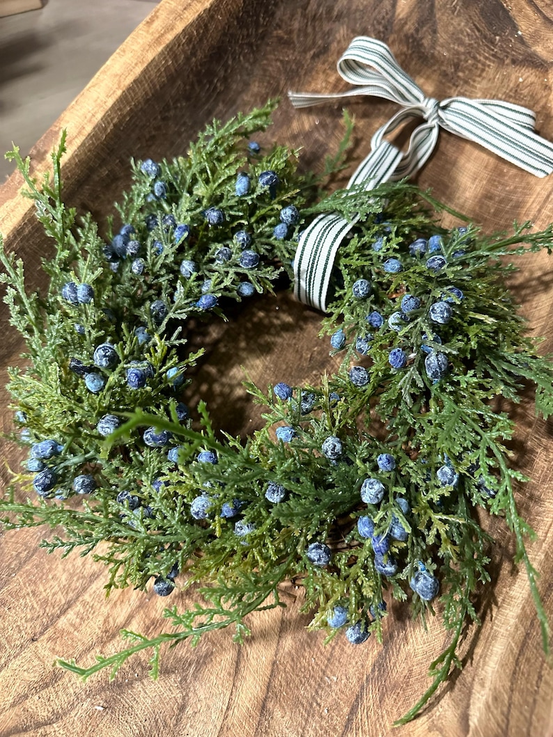 May include: A green wreath with blue berries and a white and black striped ribbon. The wreath is sitting in a wooden bowl.