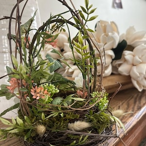 May include: A brown twig nest with green moss, small white flowers, and a green egg inside. The nest is surrounded by green leaves and twigs.
