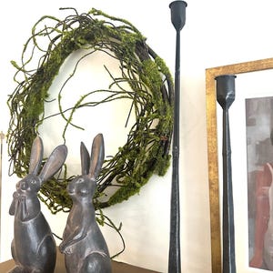 May include: Two dark gray rabbit figurines, a green moss wreath, and two tall black metal candle holders on a wooden shelf.