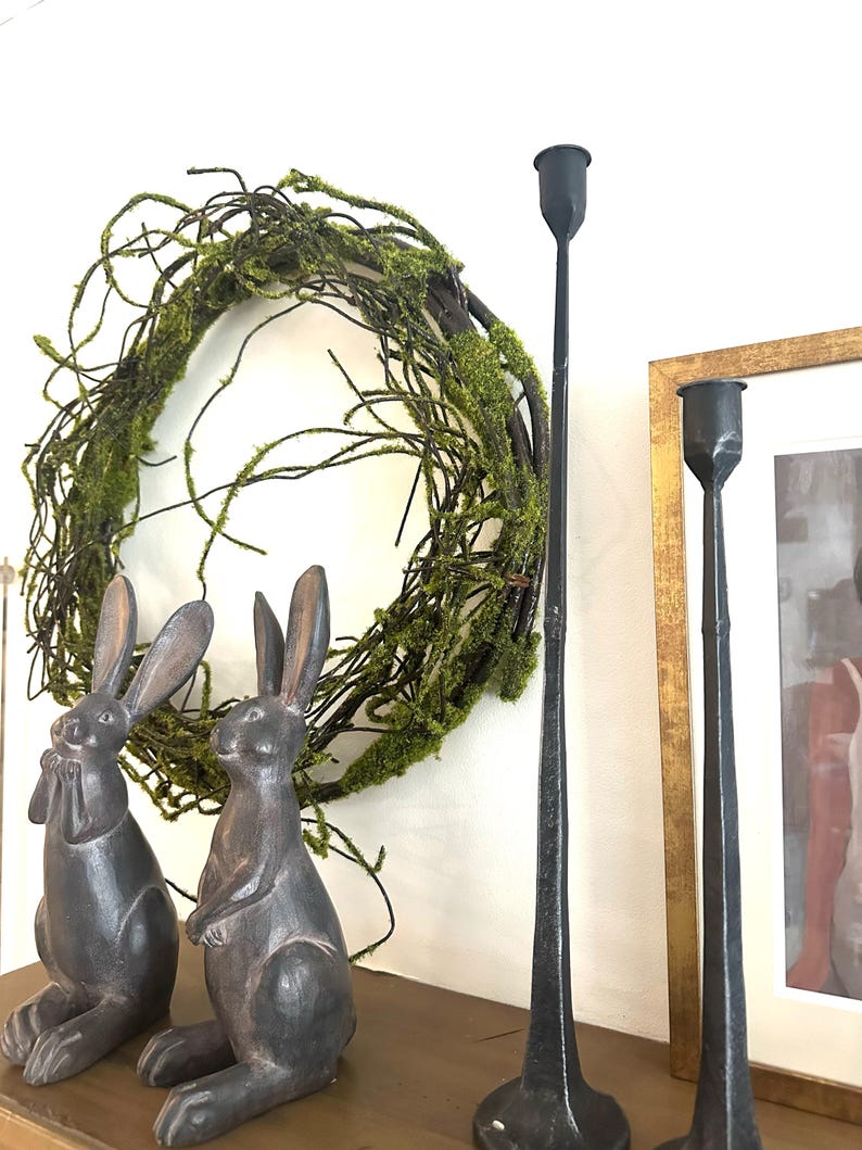 May include: Two dark gray rabbit figurines, a green moss wreath, and two tall black metal candle holders on a wooden shelf.