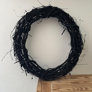 May include: A circular wreath constructed from thin, intertwined twigs and branches, entirely finished in a matte black color. The structure features a dense, organic arrangement of woody stems with various protruding ends, creating a textured, open-center ring design.