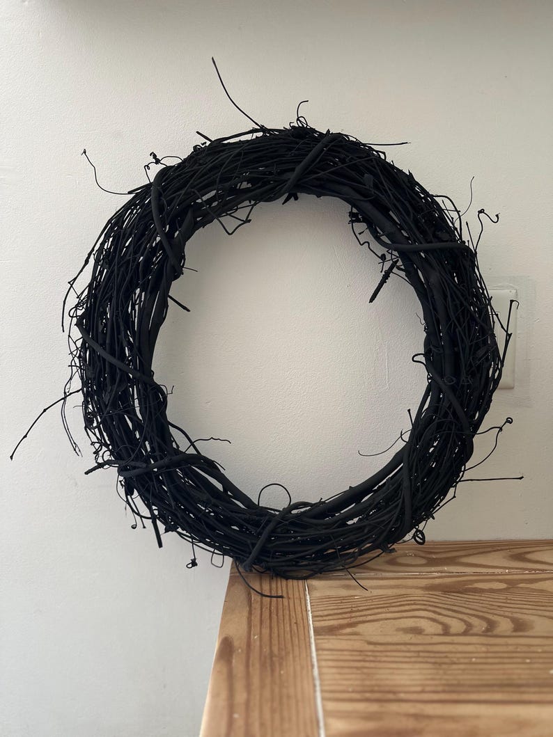 May include: A circular wreath constructed from thin, intertwined twigs and branches, entirely finished in a matte black color. The structure features a dense, organic arrangement of woody stems with various protruding ends, creating a textured, open-center ring design.