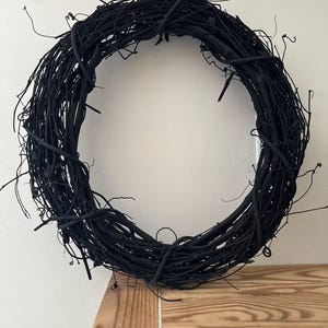 May include: A circular wreath constructed from thin, intertwined twigs and branches, entirely coated in a matte black finish. The structure features a dense, textured arrangement of organic elements with various protruding stems and thin, wiry tendrils.
