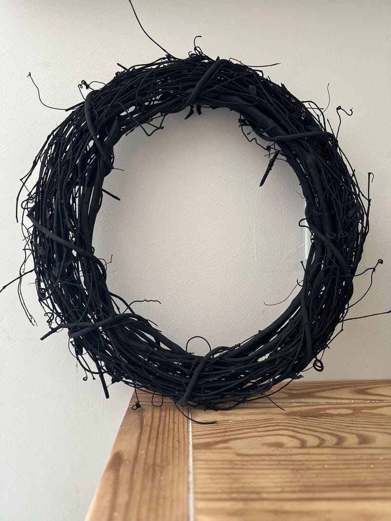 May include: A circular wreath constructed from thin, intertwined twigs and branches, entirely coated in a matte black finish. The structure features a dense, textured arrangement of organic elements with various protruding stems and thin, wiry tendrils.