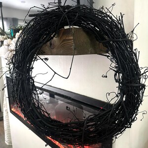 May include: A circular wreath constructed from thin, intertwined black twigs and branches. The structure features a dense, textured arrangement of dark, organic material with numerous small, protruding stems and curled tendrils throughout the frame.