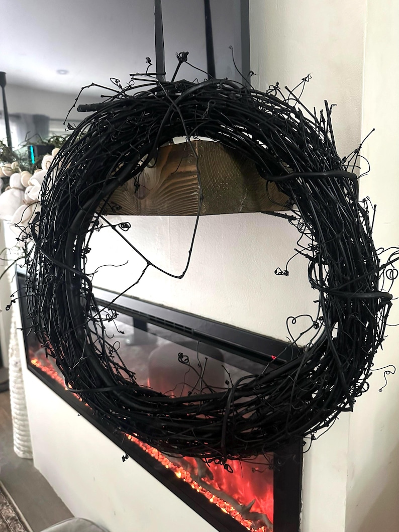 May include: A circular wreath constructed from thin, intertwined black twigs and branches. The structure features a dense, textured arrangement of dark, organic material with numerous small, protruding stems and curled tendrils throughout the frame.