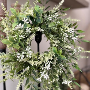May include: A green and white artificial wreath with small white flowers and green leaves. The wreath is hanging on a black metal stand.
