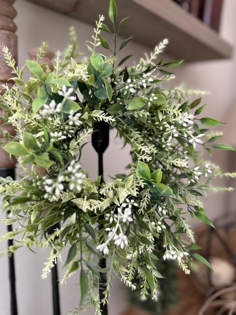 May include: A green and white artificial wreath with small white flowers and green leaves. The wreath is hanging on a black metal stand.