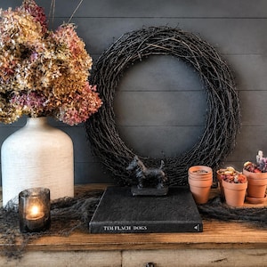 May include: A white ceramic vase filled with dried pink and brown hydrangeas, a black twig wreath, a black book titled TIM FLACH DOGS, a small black dog figurine, three terracotta pots with dried flowers, and two glass votive candle holders.