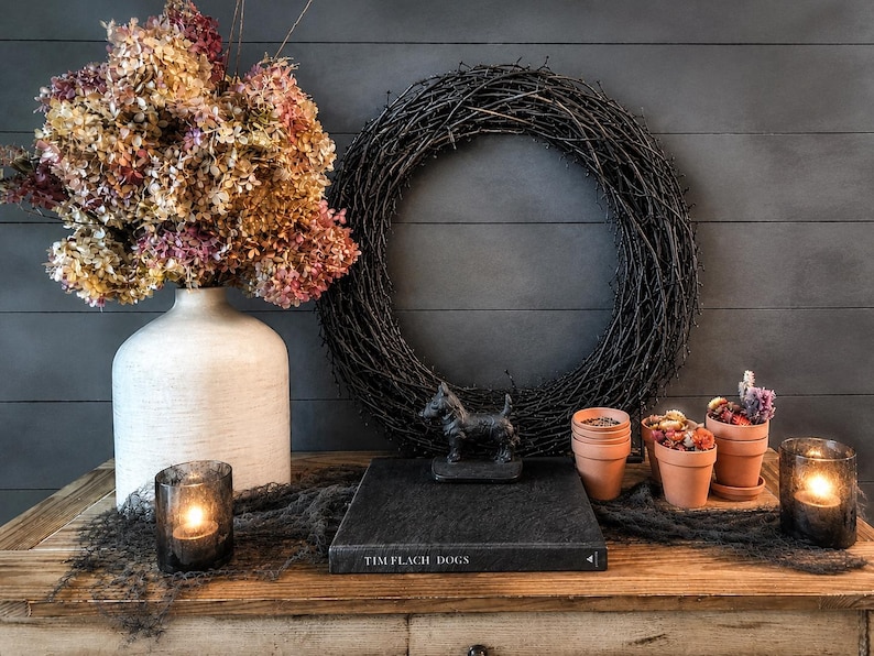 May include: A white ceramic vase filled with dried pink and brown hydrangeas, a black twig wreath, a black book titled TIM FLACH DOGS, a small black dog figurine, three terracotta pots with dried flowers, and two glass votive candle holders.