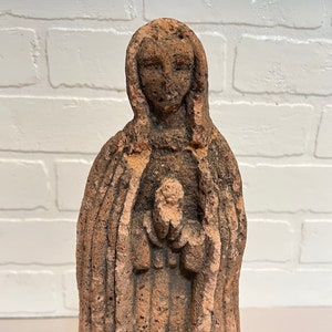 Puede incluir: Una estatua de terracota de una mujer con una larga túnica, sosteniendo una pequeña figura en sus brazos. La estatua está desgastada y tiene una textura rugosa.
