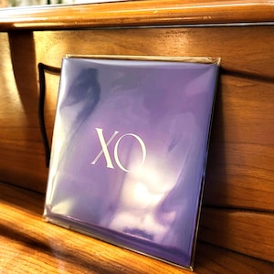 May include: A square, purple item in a clear plastic sleeve. The text "XO" is printed in white on the front. The item is resting on a wooden surface.