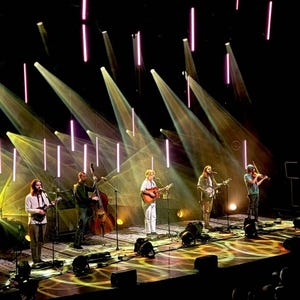 May include: A band of five musicians performing on stage with microphones and instruments, including a guitar, banjo, and violin. The stage is lit with spotlights and has a patterned floor.