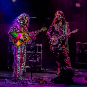 Puede incluir: Dos músicos tocando en un escenario. Un músico lleva un traje de pantalón rosa y blanco a rayas y toca una guitarra acústica marrón. El otro músico lleva una chaqueta de lentejuelas moradas y toca un banjo.